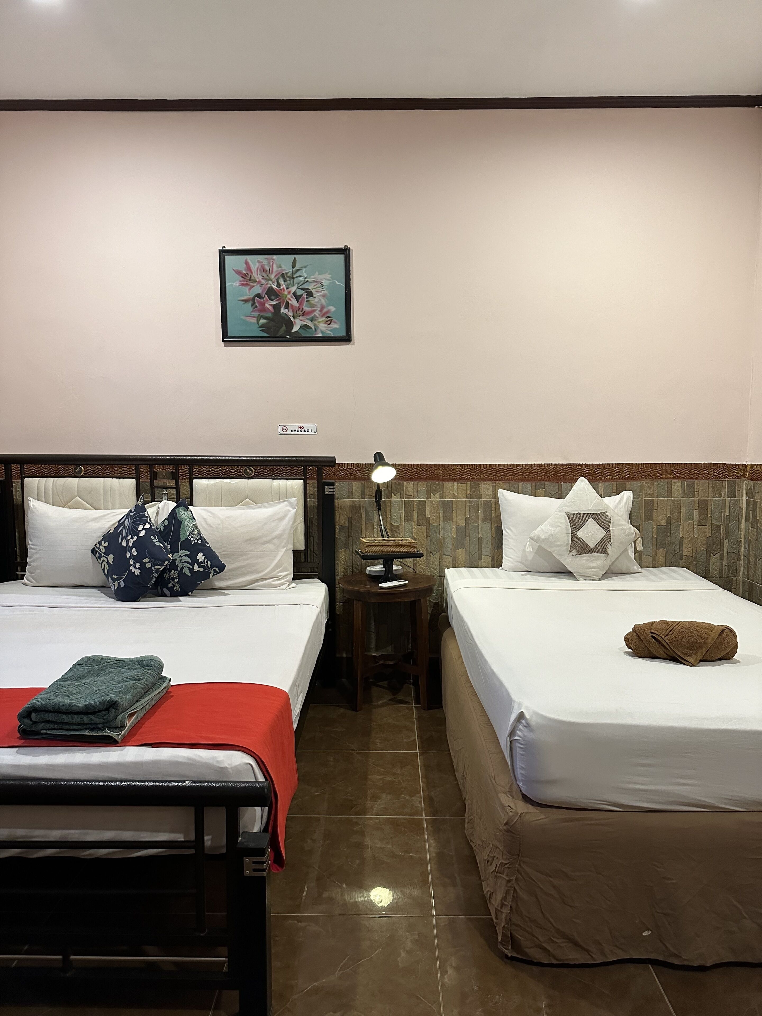 A hotel room with two beds, a small table with a lamp, a painting on the wall, and a 'No Smoking' sign.