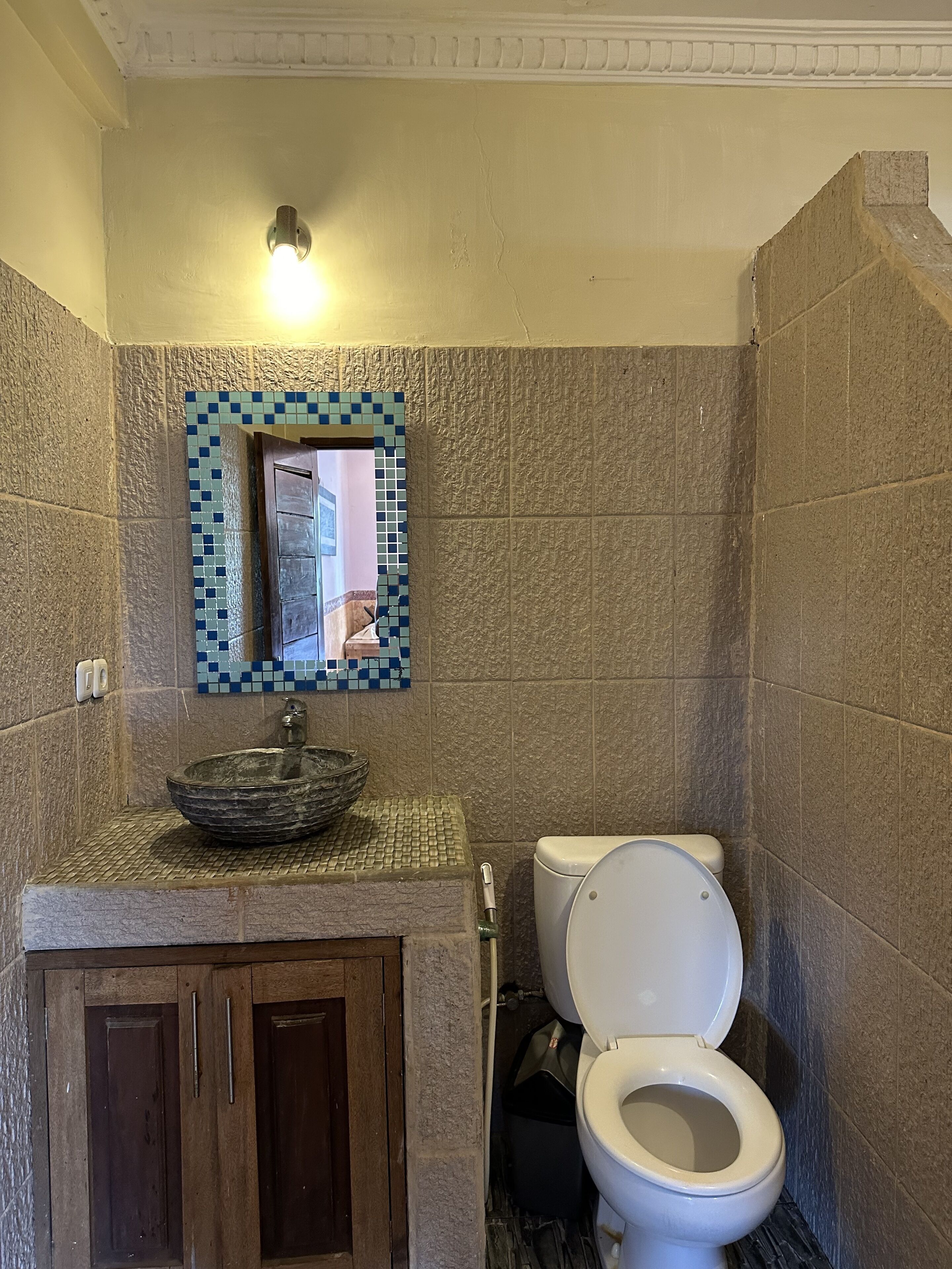 A bathroom with a mosaic tile wall, a glass bowl sink, a white toilet, and wooden cabinets.