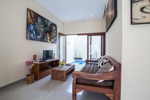 A modern living room with a sofa, coffee table, and TV stand. There is a large painting on the wall and a window with a view of the outdoors. - Villa D'Kerobokan (Denpasar)