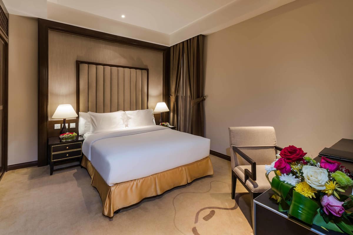 Habitación de hotel con una cama grande, dos mesitas de noche con lámparas, una silla y un escritorio con una computadora portátil y flores.