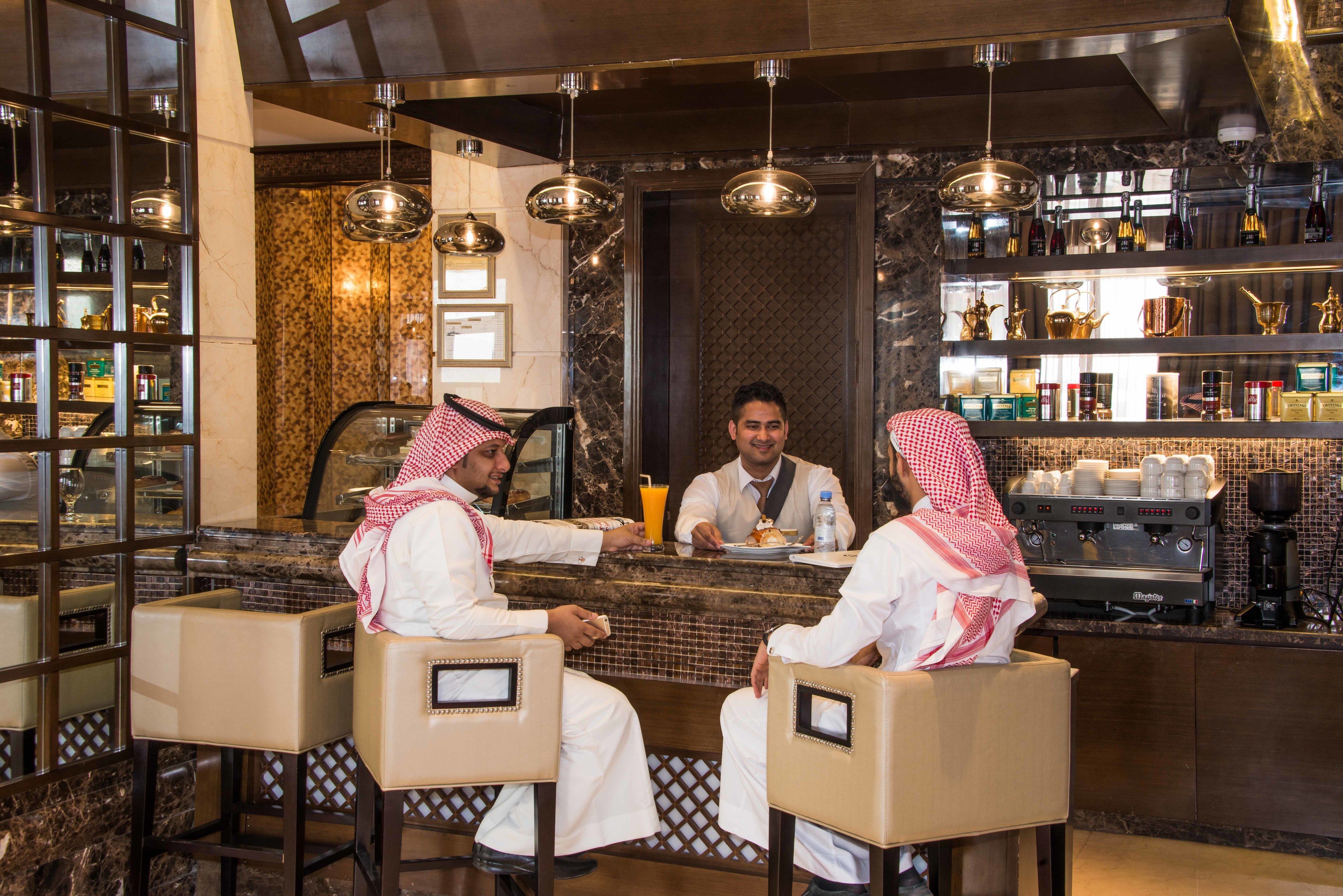 A bar area with three seated individuals, a counter with a coffee machine, and shelves with bottles and glasses.