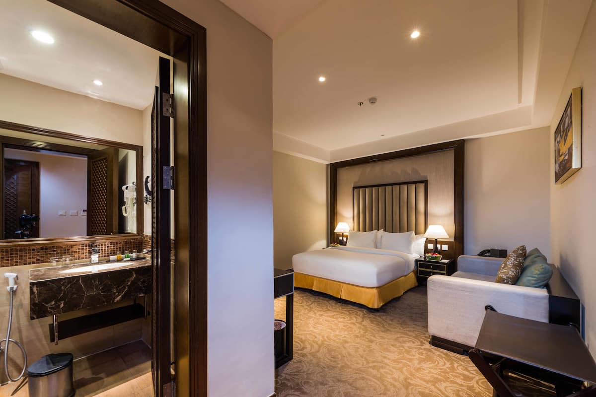 Habitación de hotel con cama, mesita de noche, lámpara y baño con lavabo y espejo.
