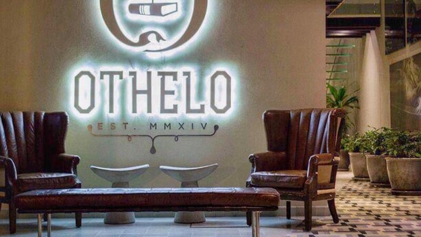 Un vestíbulo con un letrero de neón que dice "OTHELO EST. MMXIV" y dos sillones de cuero frente a una mesita.