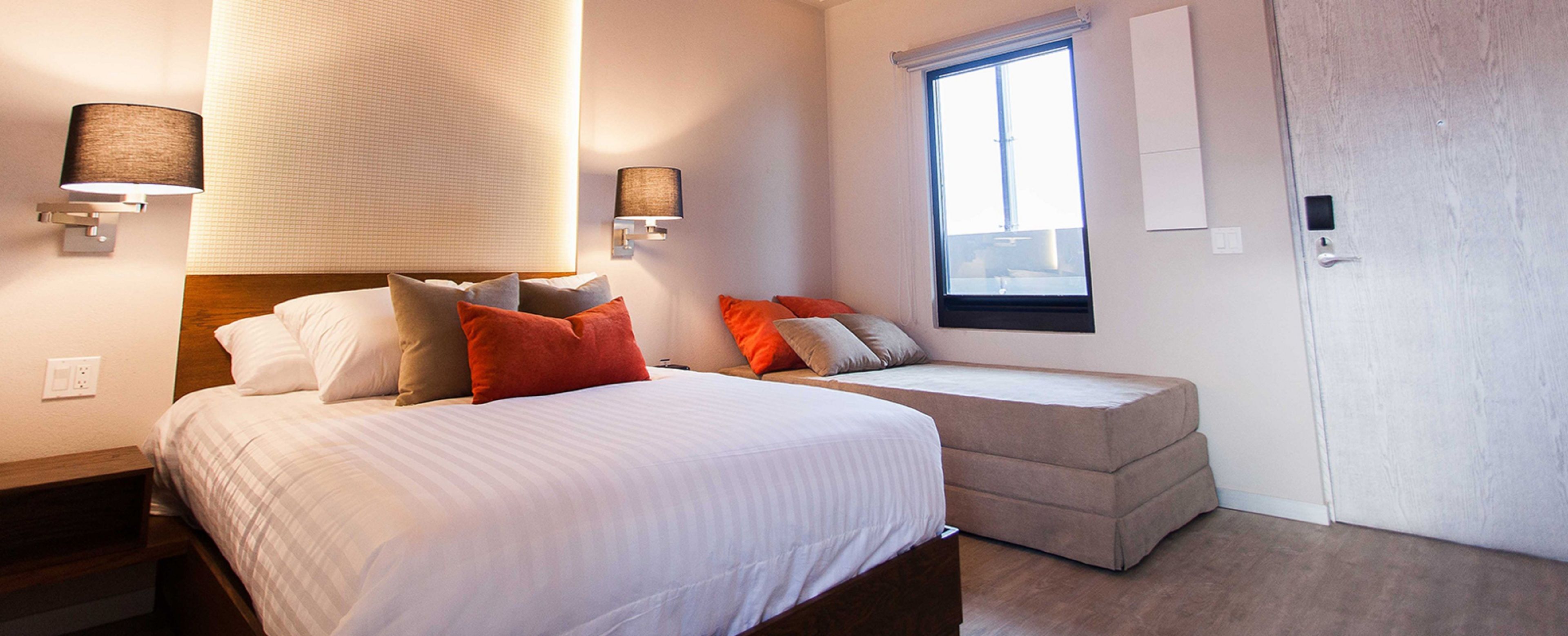 A bedroom with a bed, a sofa bed, two wall-mounted lamps, a window, and a door.