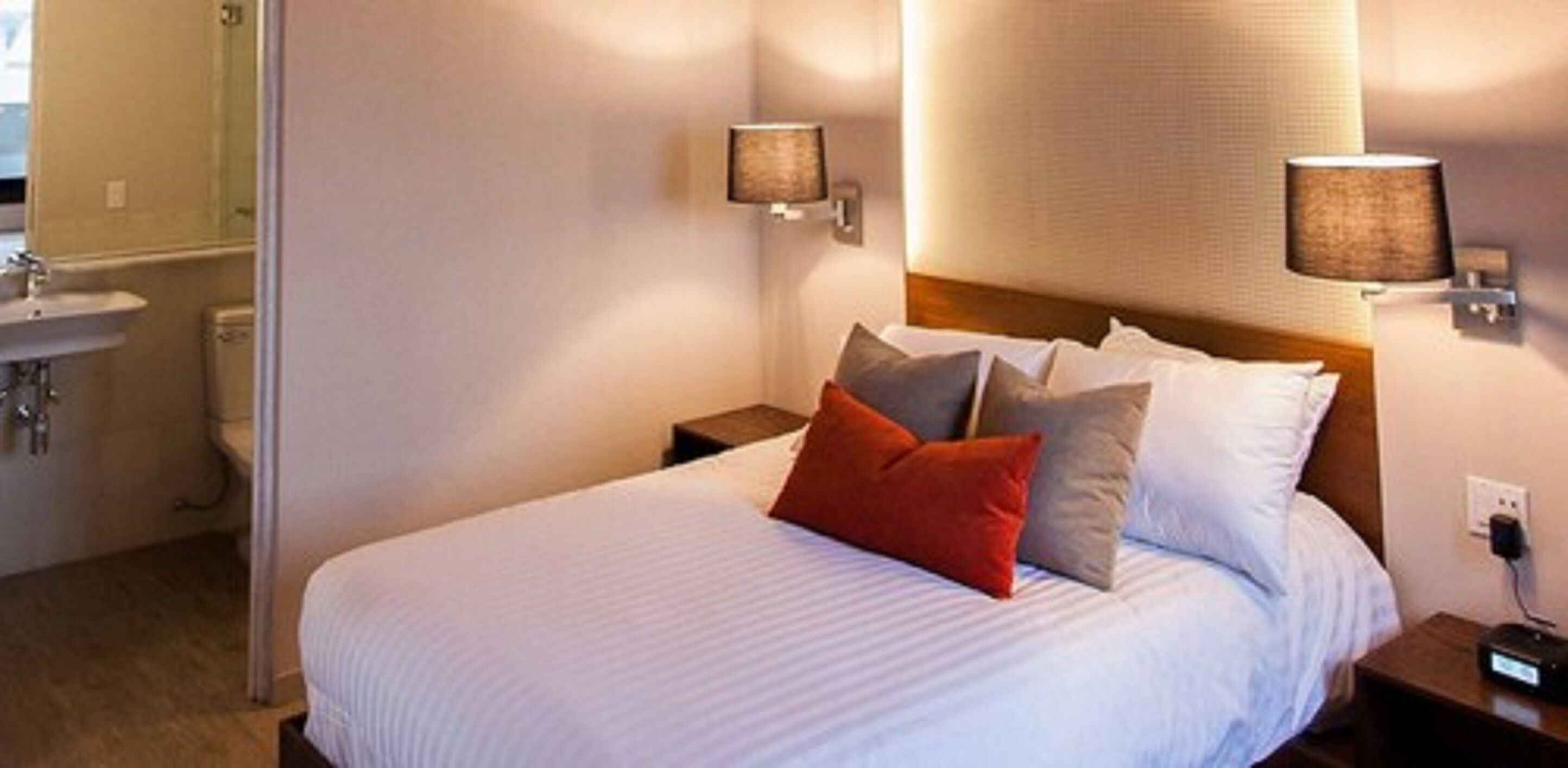 Habitación de hotel con una cama, mesitas de noche, una lámpara de pared y un baño visible al fondo.