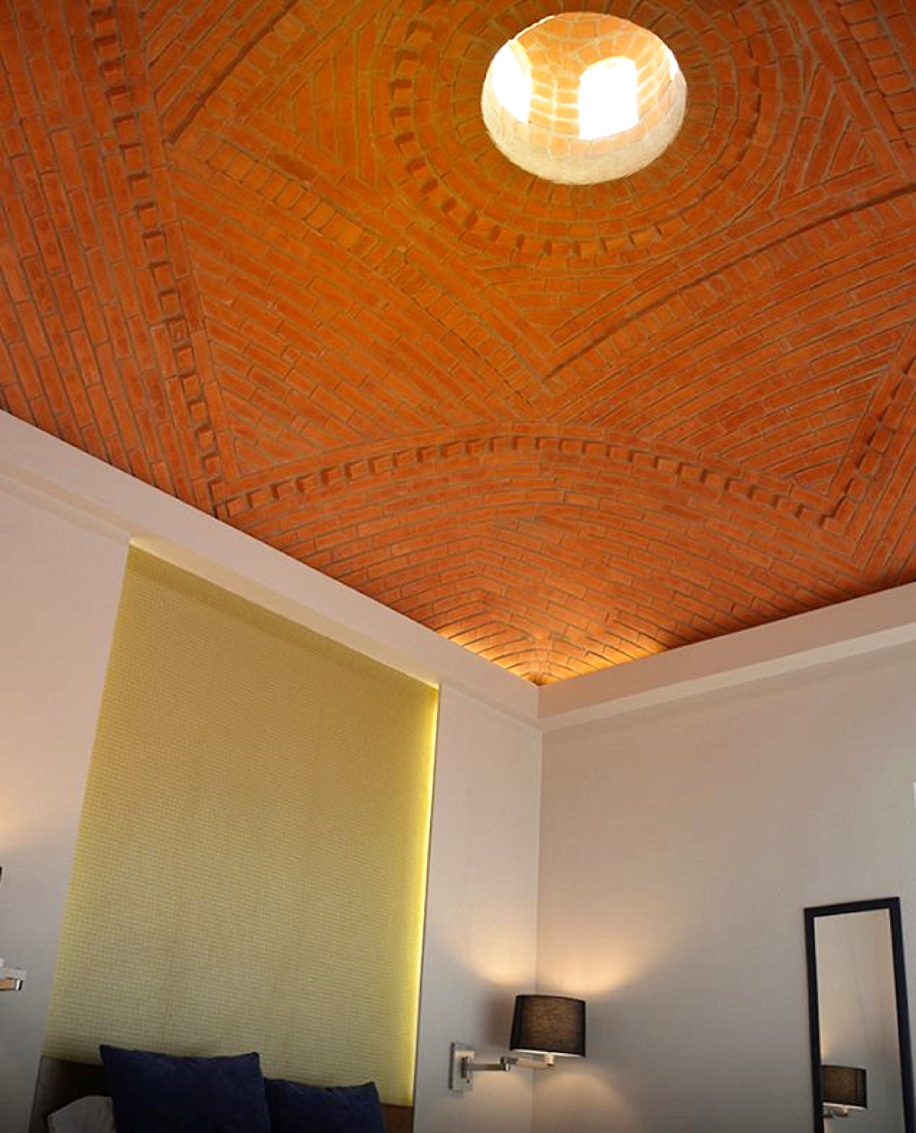 A room with a brick-patterned ceiling, a bed with pillows, a wall-mounted lamp, and a mirror.