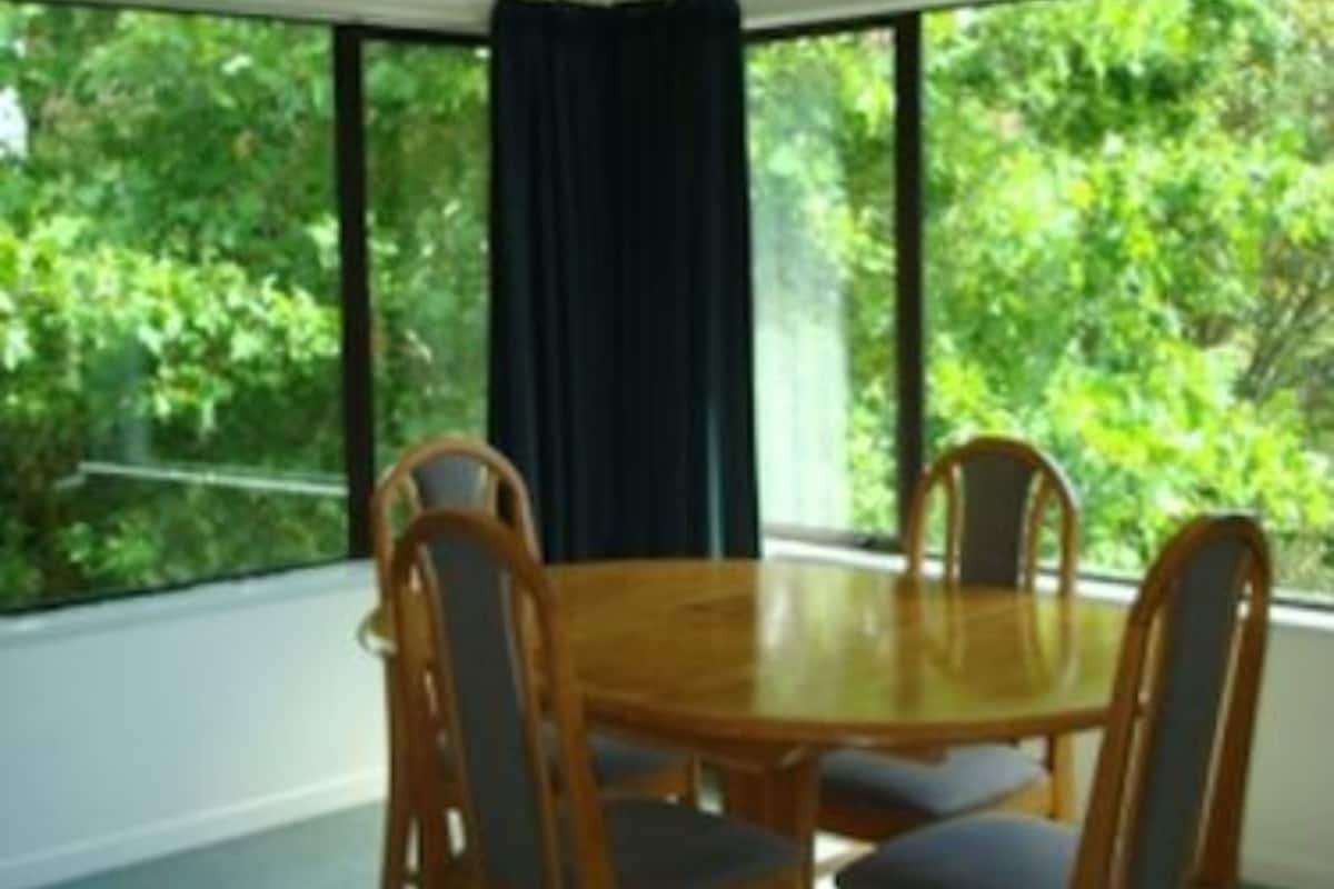 Un coin repas avec une table ronde et quatre chaises, de grandes fenêtres donnant sur un paysage verdoyant et un rideau foncé.