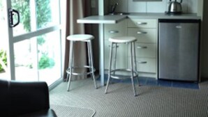 Une cuisine moderne avec des meubles blancs, un réfrigérateur en acier inoxydable et une bouilloire posée sur le plan de travail. On y trouve deux tabourets blancs et un comptoir-bar.