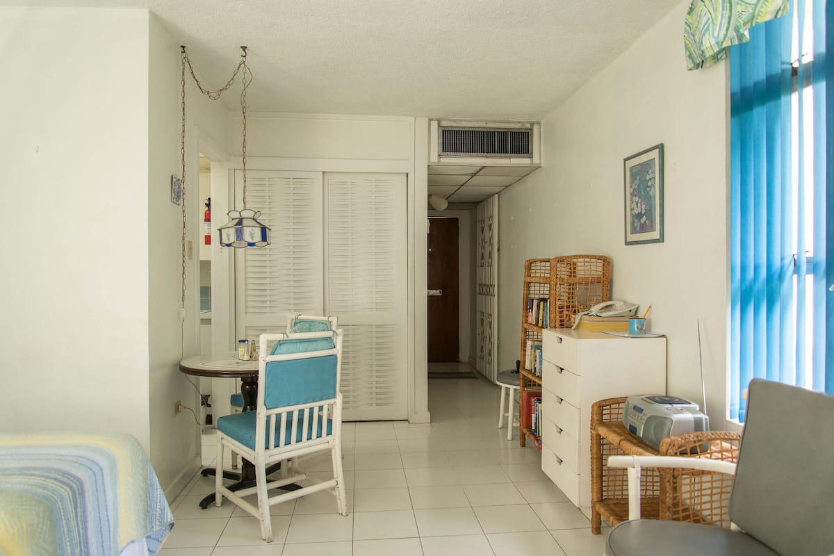 Una habitación pequeña y bien iluminada con una cama, un escritorio con una silla, una estantería y una silla de mimbre.