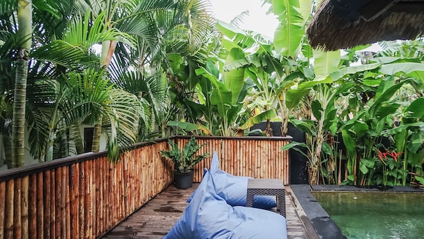 A wooden deck with blue bean bags, surrounded by lush tropical plants and a pool. - Kampung 168 (Jimbaran)