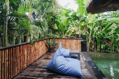 A wooden deck with blue bean bags, surrounded by lush tropical plants and a pool.