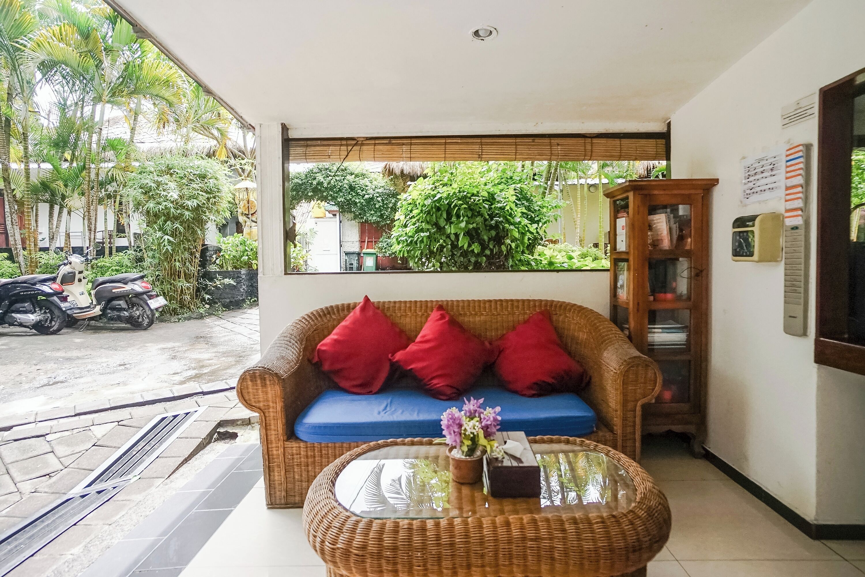 Una habitación con un sofá de mimbre, cojines rojos, una mesa de centro de vidrio con un florero y vistas a la calle y al verdor exterior.