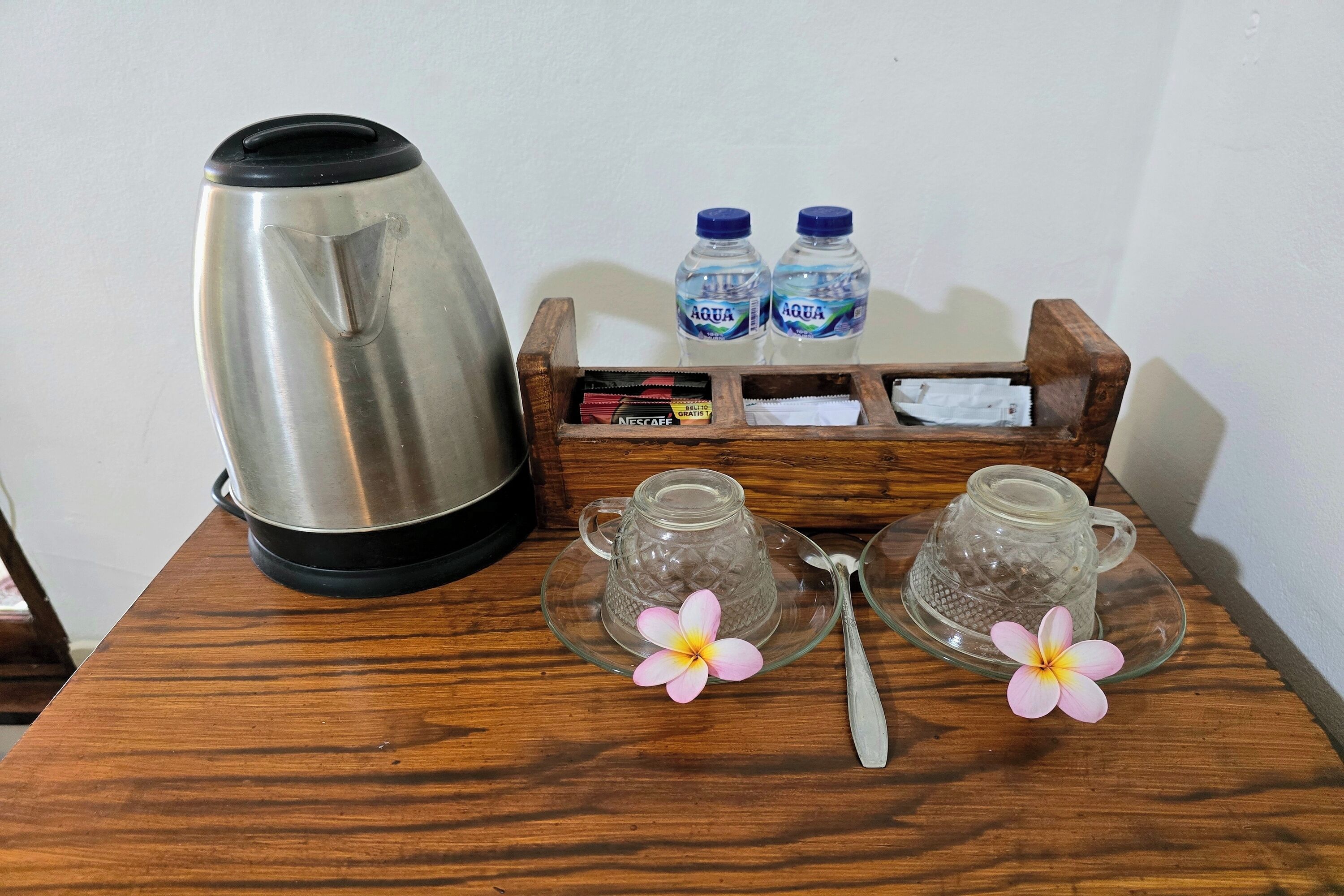 Una mesa de madera con una tetera de acero inoxidable, dos tazas de vidrio, dos botellas de agua, un paquete de café Nescafé y dos adornos florales.