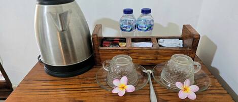 Una mesa de madera con una tetera de acero inoxidable, dos tazas de vidrio, dos botellas de agua, un paquete de café Nescafé y dos adornos florales.