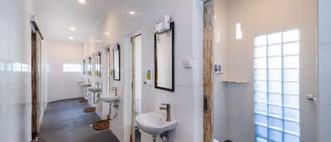 Un baño largo y estrecho con varios lavamanos y espejos, un ventanal amplio y un cesto de basura.