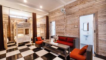 Quarto de hotel com piso xadrez, sofá com almofadas laranja, beliche de madeira e espelho na parede.