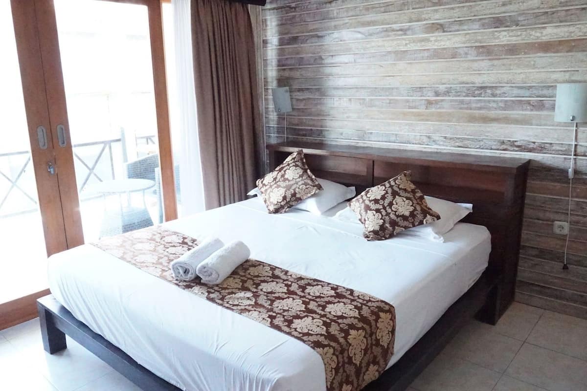 Habitación de hotel con cama, almohadas, mesita de noche, lámpara y vistas al exterior.