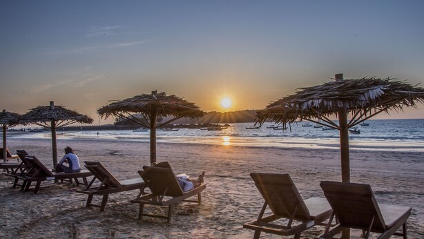 Una playa con sillas de descanso y sombrillas de paja, un atardecer y barcos en el agua.