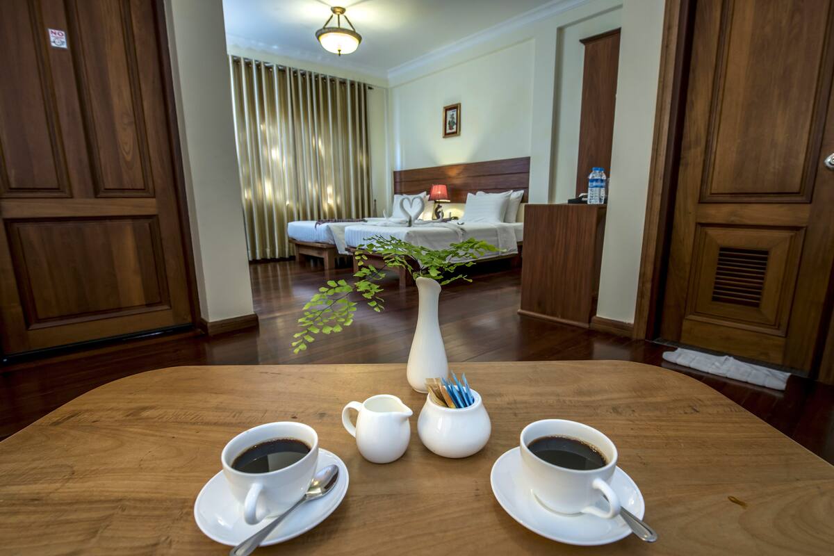 A hotel room with a wooden floor, a bed with white bedding, a bedside table with a lamp, and a vase with a plant. There are two cups of coffee on the table.