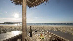 Escena de playa con una mesa preparada para dos bajo un techo de paja, una botella de vino y un plato de comida.