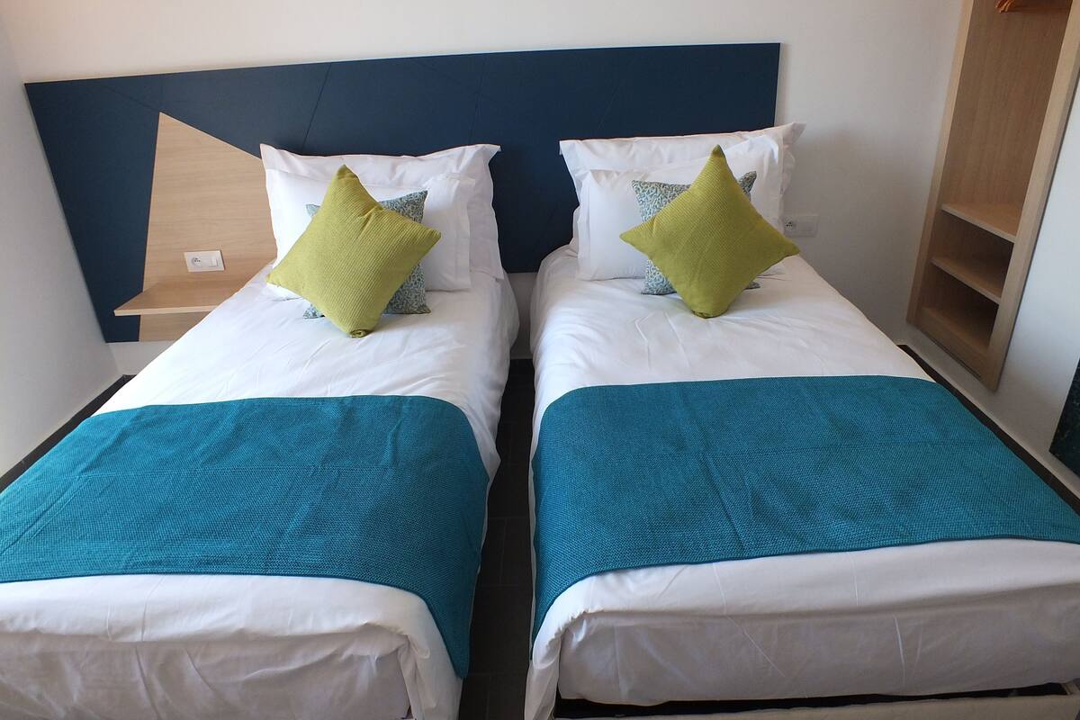 A double bed with white and teal bedding, a blue headboard, and a wooden nightstand with a small shelf.