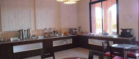 A breakfast bar with coffee machines, a buffet with various food items, and seating with red cushioned chairs.