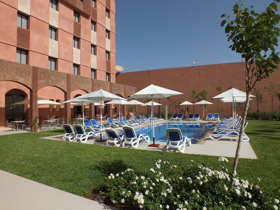 A poolside area with lounge chairs and umbrellas, a swimming pool, and a building with brick arches and a flat roof.