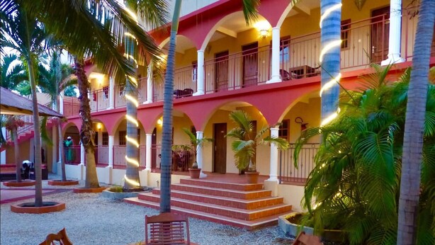 Un patio con un edificio rojo y amarillo, palmeras y mesas al aire libre.