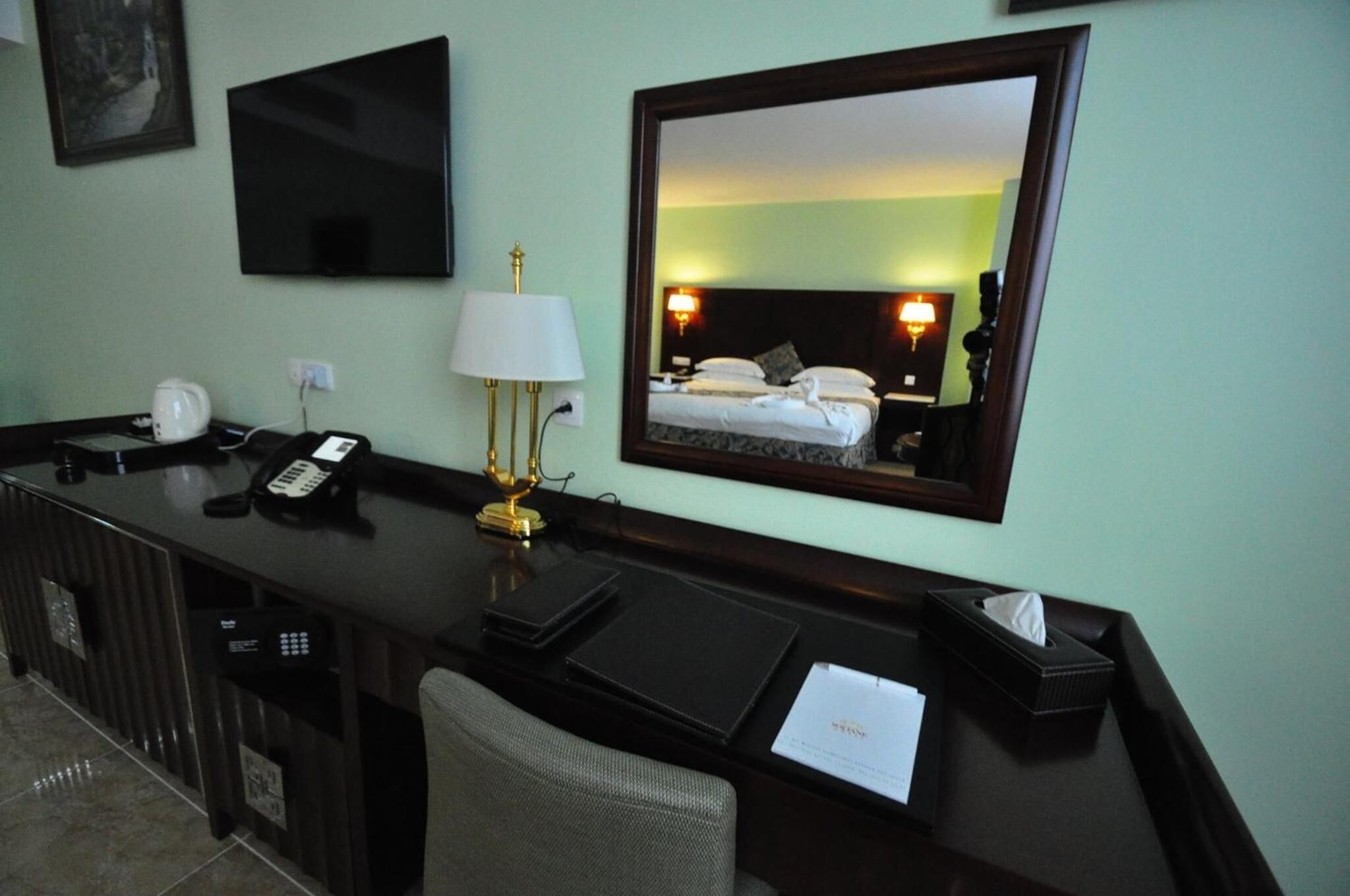 A hotel room with a desk, a telephone, a lamp, a mirror, and a bed with a headboard.
