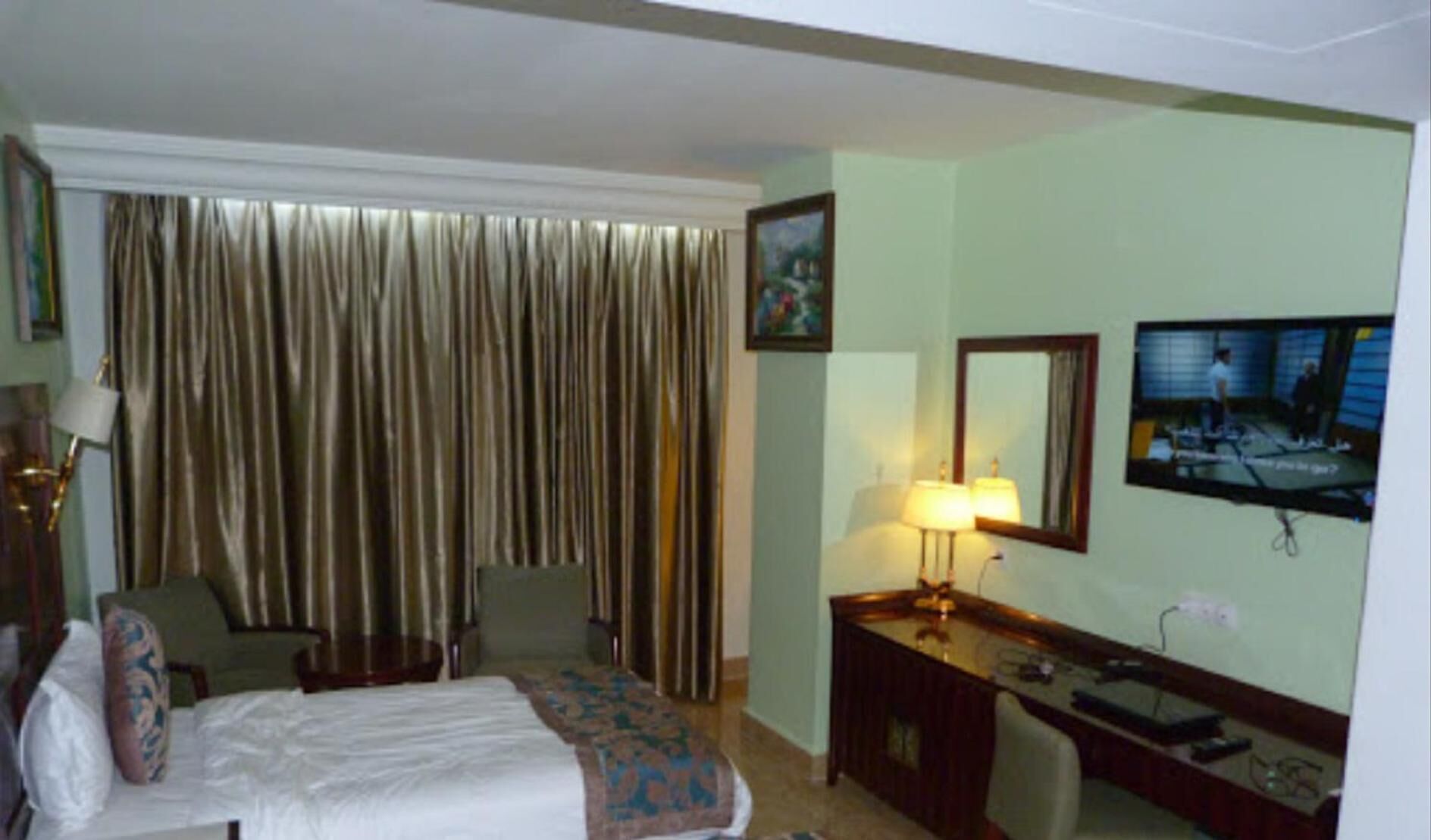 Ein Hotelzimmer mit einem Bett, einem Schreibtisch, einem Sessel, einer Lampe, einem Fernseher und einem großen Fenster mit Vorhängen.