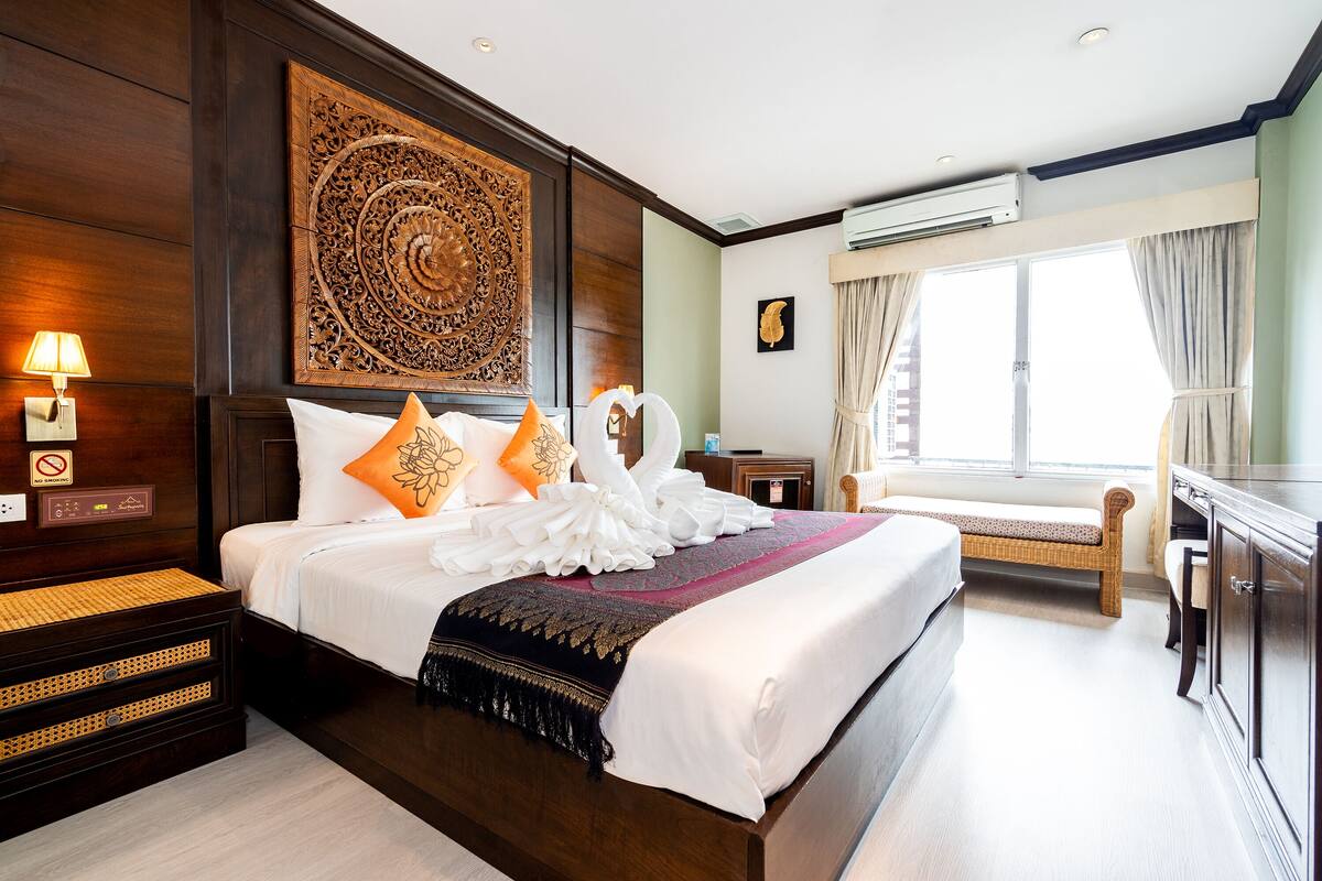 Habitación de hotel con una cama grande, cabecera de madera, aire acondicionado de pared, mesita de noche con lámpara y ventana con cortinas.