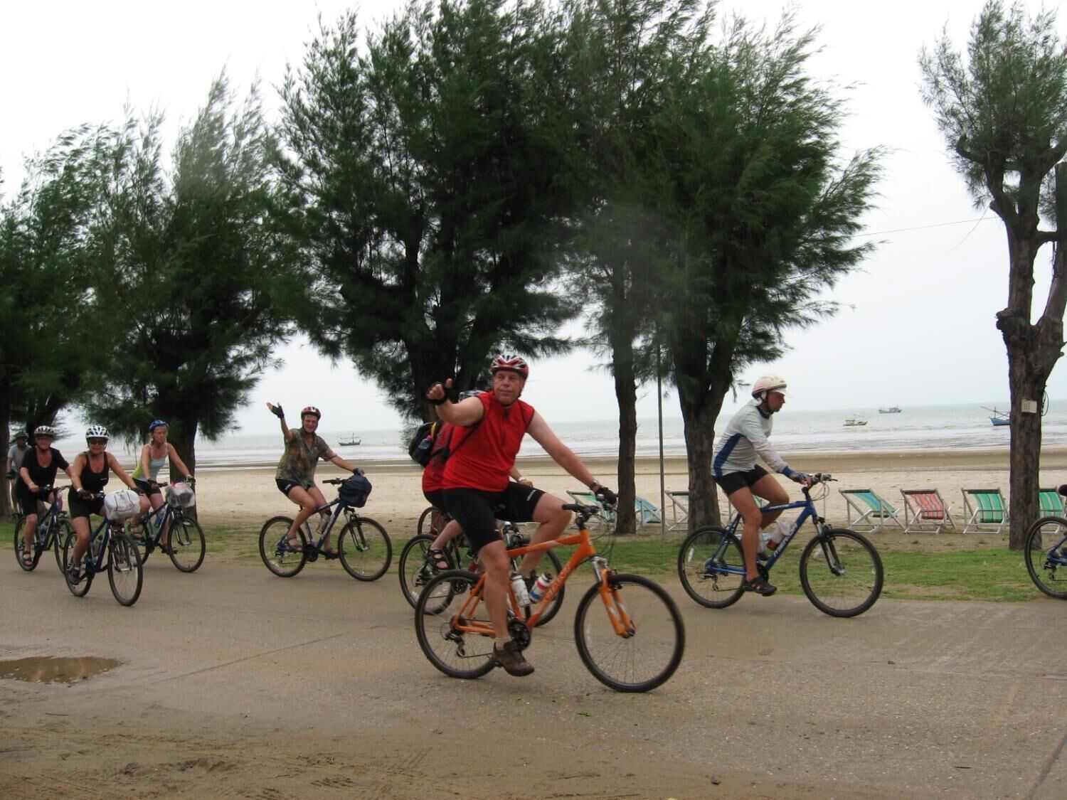 Un grupo de ciclistas en un camino pavimentado cerca de una playa, con árboles y sillas de playa al fondo.