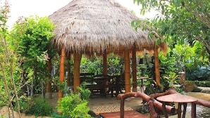 A thatched-roof gazebo with a wooden bench inside, surrounded by lush greenery and a small pond.