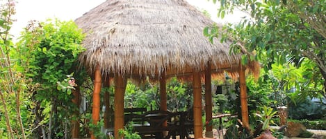 Un belvédère au toit de chaume, avec un banc en bois à l’intérieur, entouré d’une végétation luxuriante et d’un petit étang.