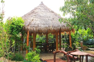A thatched-roof gazebo with a wooden bench inside, surrounded by lush greenery and a small pond.