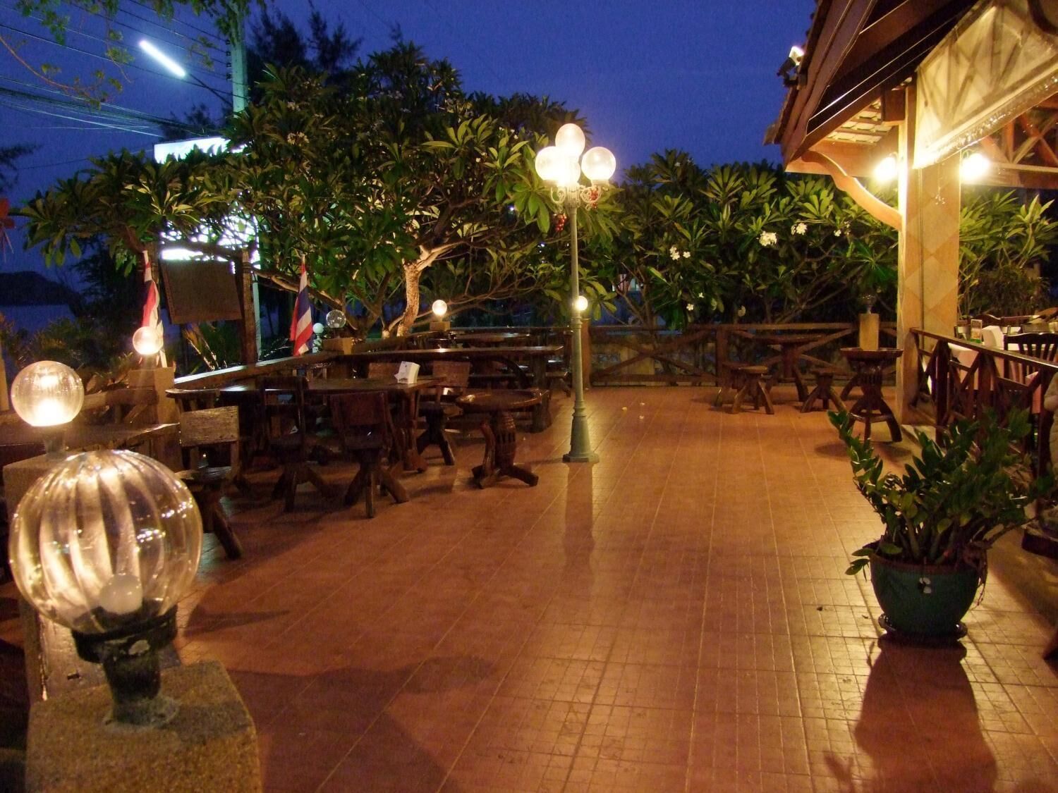 An outdoor dining area with wooden tables and chairs, surrounded by potted plants and trees.