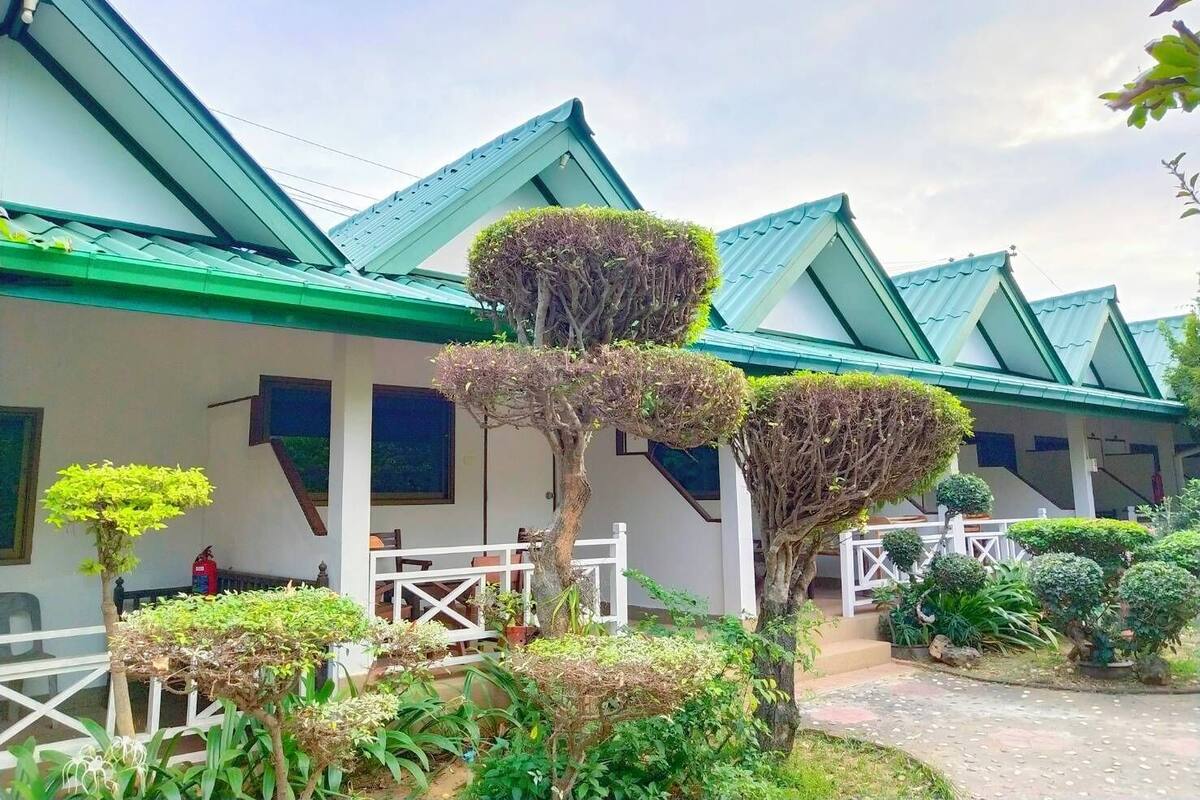 Um edifício com telhado verde e paredes brancas, cercado por diversas plantas e árvores.