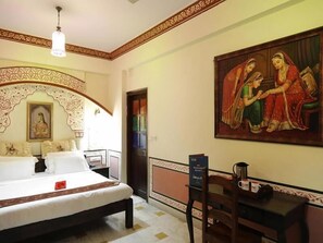 A hotel room with a bed, a desk, a chair, a painting, and a door. - OYO Rooms Heritage Hawa Sadak (Jaipur)