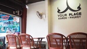 A restaurant interior with wicker chairs, a table, a wall sign reading "Your Place Karon · Phuket", and a booking center sign. - Your Place Inn (Karon)