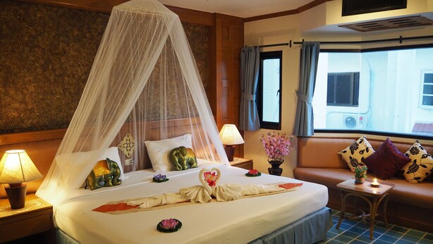 A hotel room with a bed, a nightstand, a bedside table, a window, and a view of the outside. - Your Place Inn (Karon)