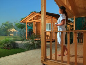 Eine Holzterrasse mit einer Person in einer weißen Robe, die eine Tasse hält und auf ein Holzgebäude sowie eine üppige grüne Landschaft blickt.