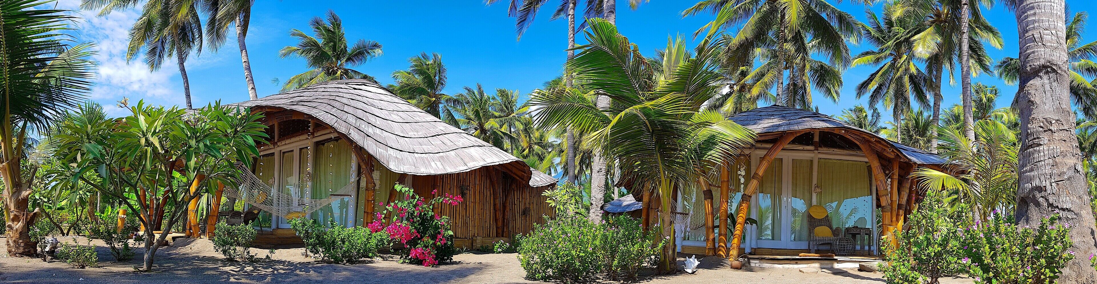 A tropical resort with two wooden huts surrounded by palm trees and lush greenery.