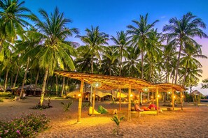 A beach with palm trees, a wooden gazebo, and thatched-roof huts.