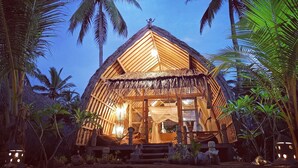 Een rieten hut met een houten constructie, omgeven door palmbomen, verlicht met binnenen verlichting.