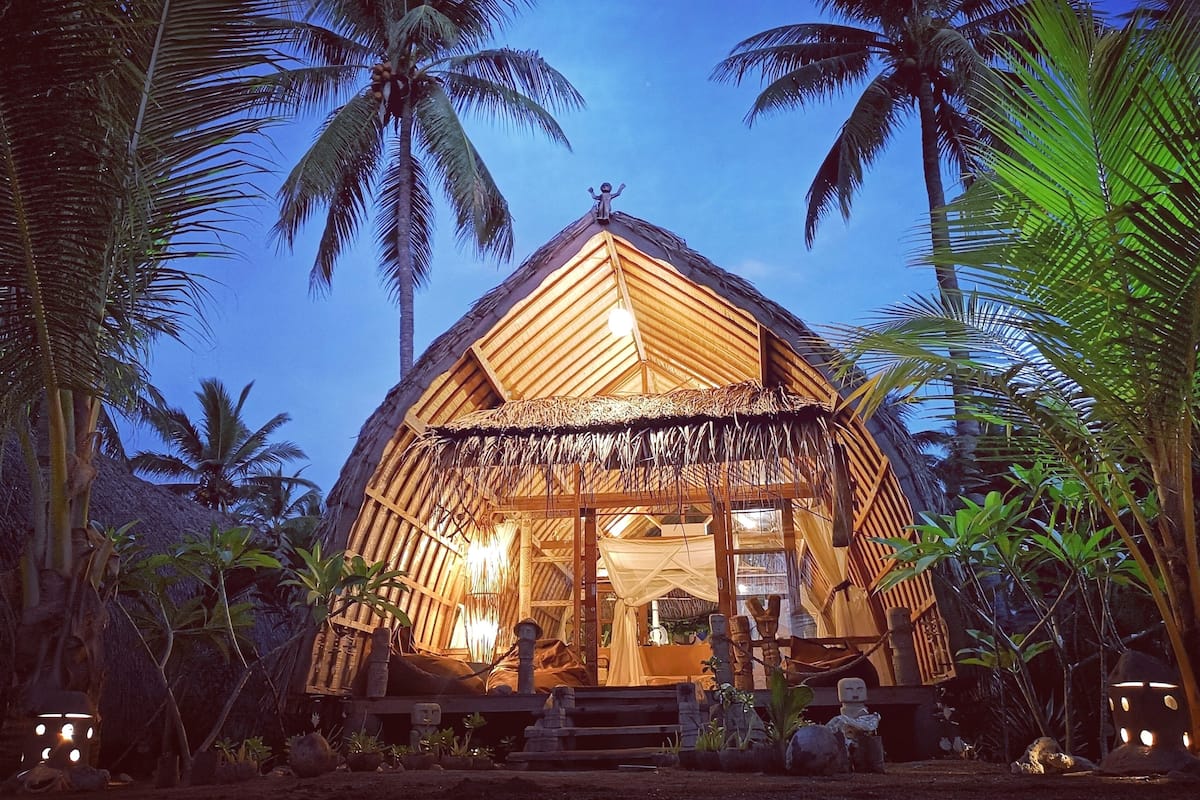 Une hutte au toit de chaume, dotée d’une structure en bois, entourée de palmiers et éclairée par un système d’éclairage intérieur.