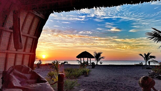 Een uitzicht op het strand met een rieten hut, palmbomen en een zonsondergang.