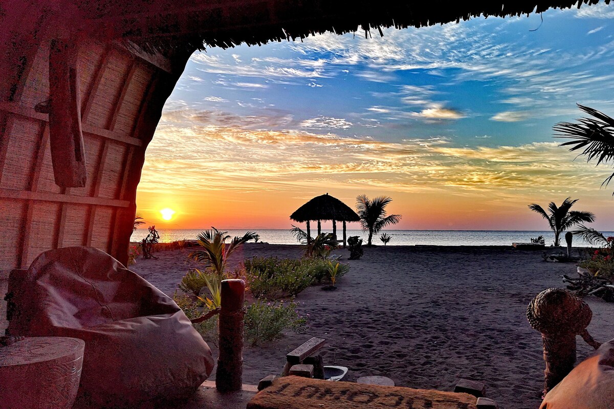 Une vue sur la plage avec une structure à toit de chaume, des palmiers et un coucher de soleil.