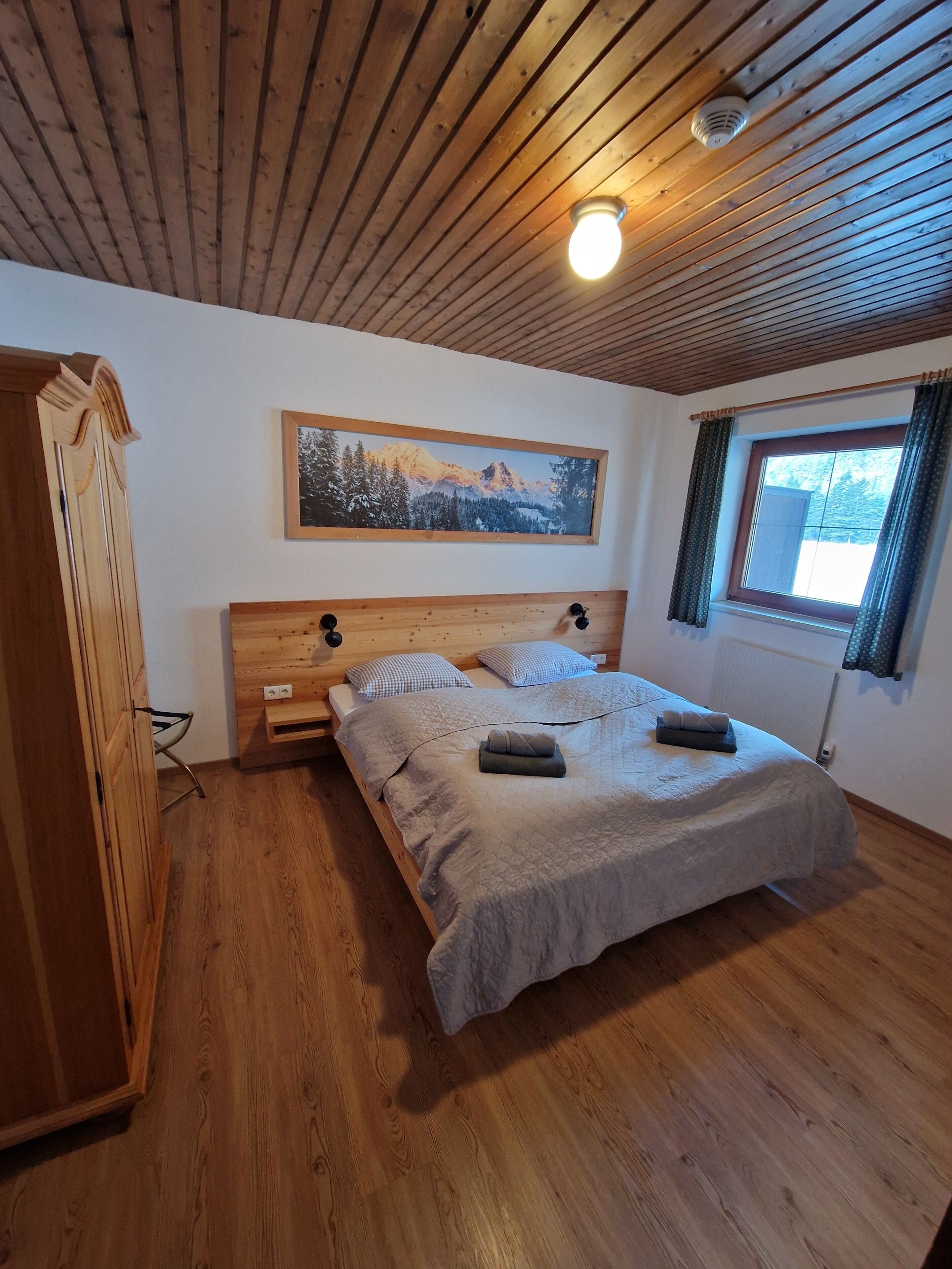 A bedroom with a wooden ceiling, a bed with a grey quilt, a wooden wardrobe, a framed picture on the wall, and a window with curtains.