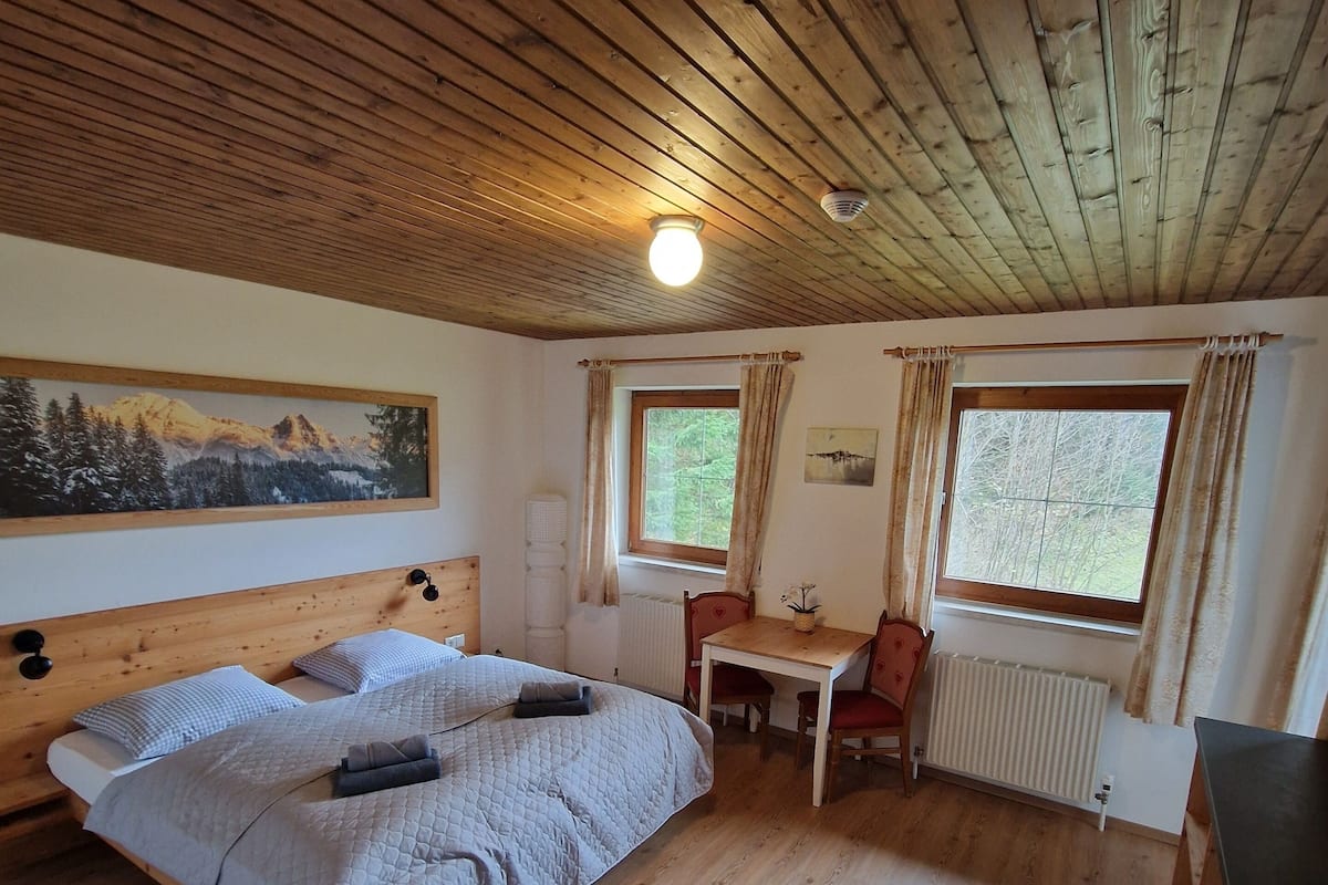 Une pièce aux murs lambrés, avec un lit, une petite table et une fenêtre avec des rideaux.
