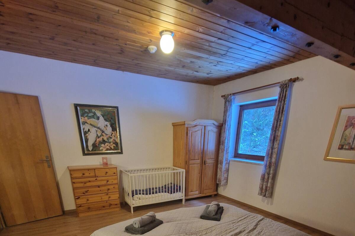 Une chambre avec un plancher en bois, un lit recouvert d’une couverture matelassée, un berceau, une armoire et une fenêtre avec des rideaux.